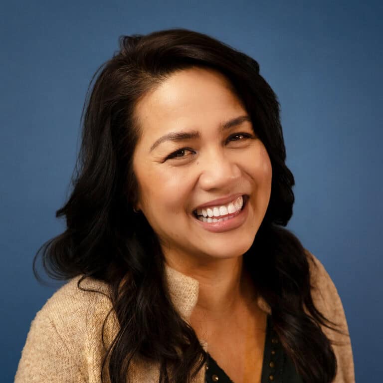 Lori Disney smiling with dark hair and a tan sweater against a blue background.
