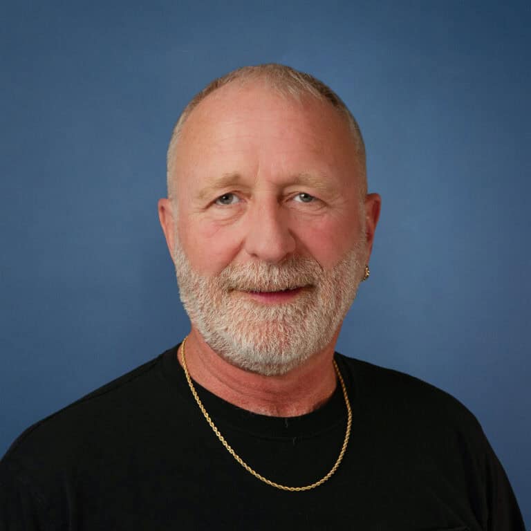 Portrait of Allen Seaman, a man with a gray beard, wearing a black shirt and gold chain against a blue background.