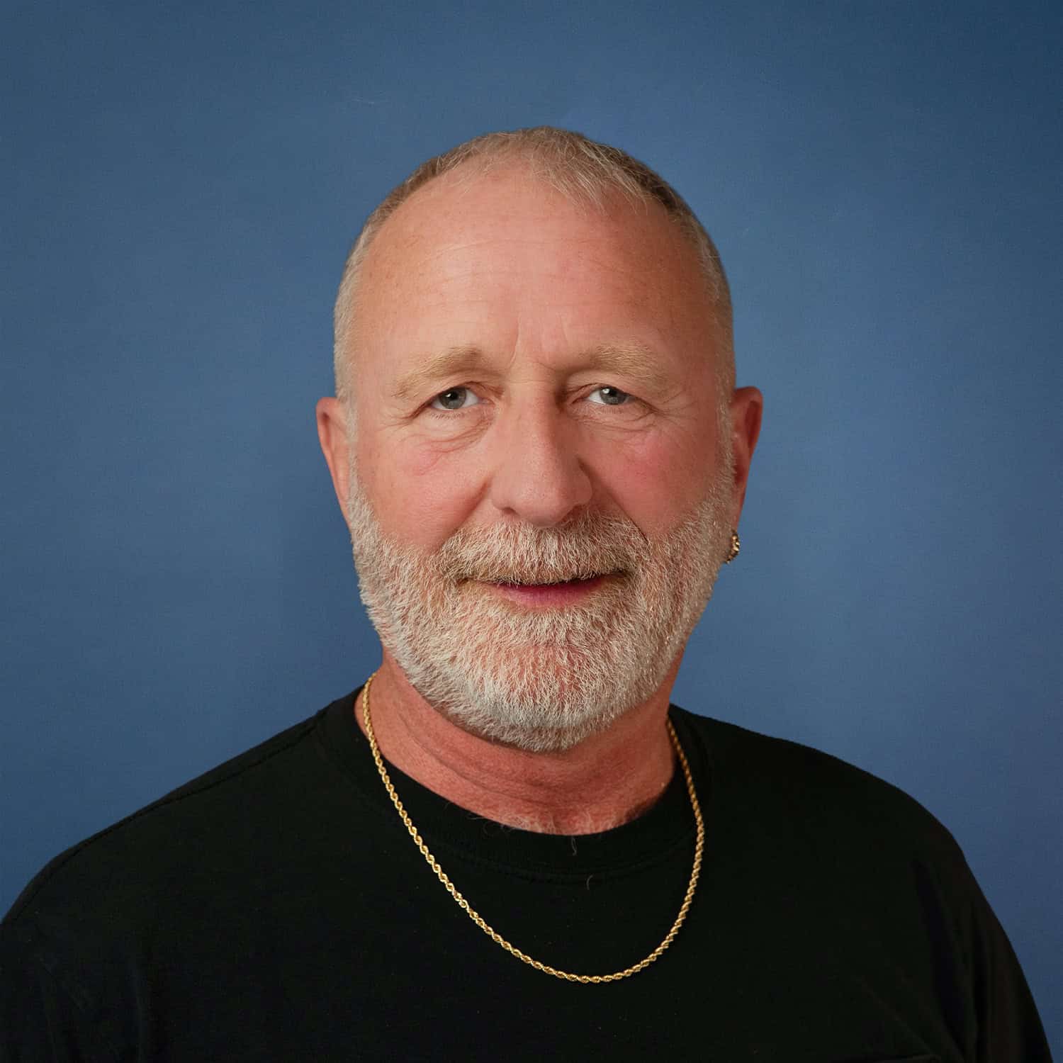 Portrait of Allen Seaman, a man with a gray beard, wearing a black shirt and gold chain against a blue background.