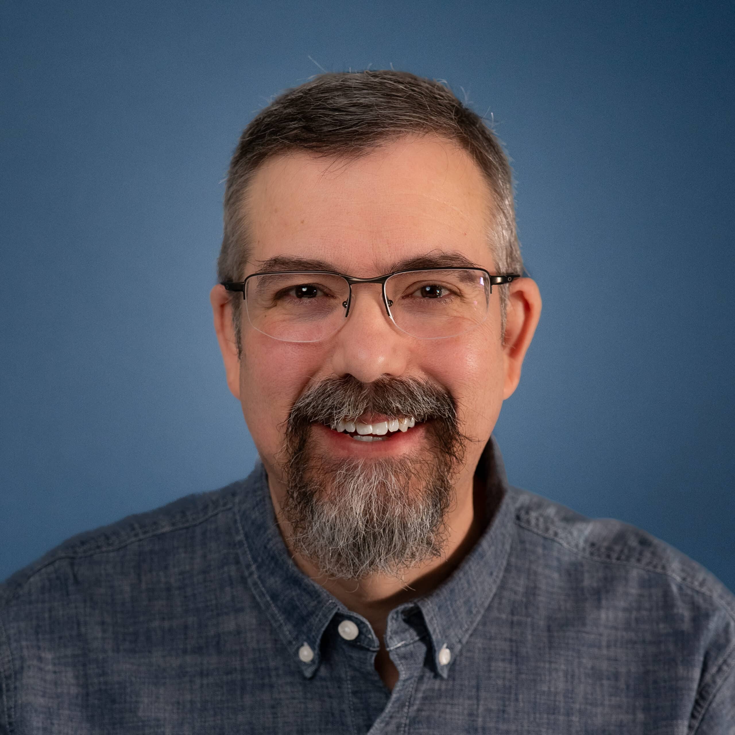 Headshot of Jason Krogmann with glasses and a beard against a blue background.