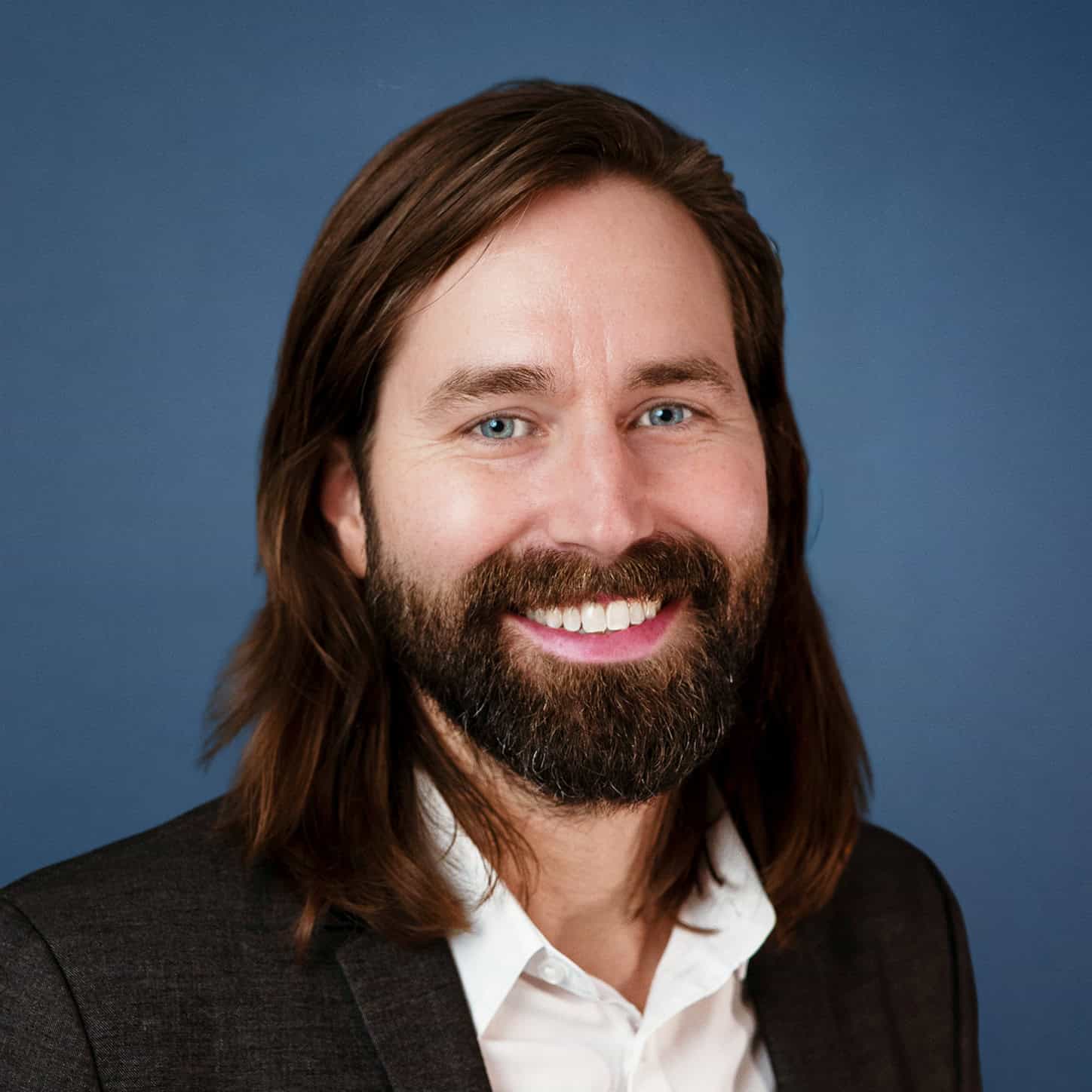 Gabe Coyle headshot, smiling with long brown hair and beard, wearing a suit.