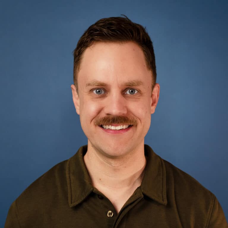 Sean Dennis portrait with a mustache, smiling against a blue background.