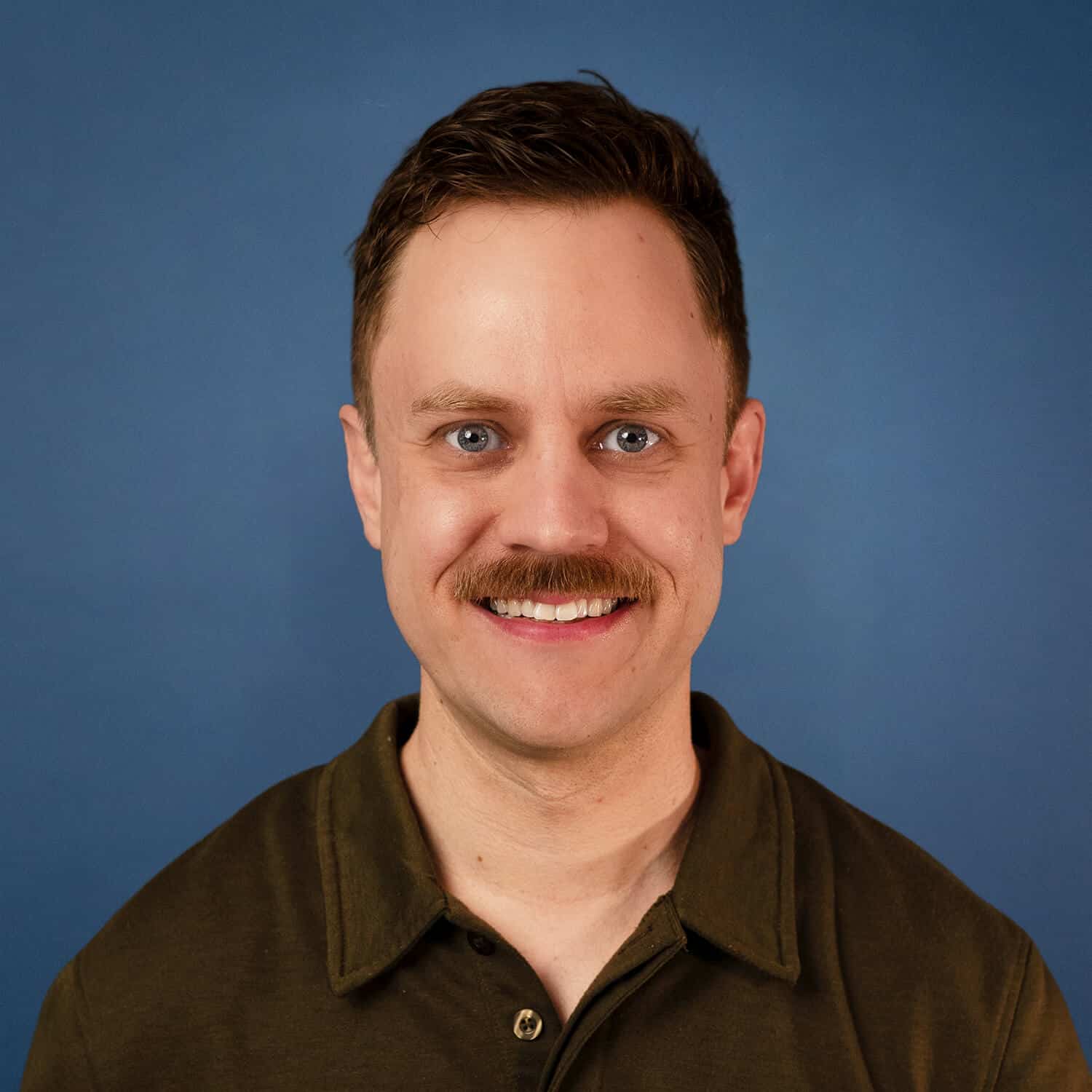 Sean Dennis portrait with a mustache, smiling against a blue background.