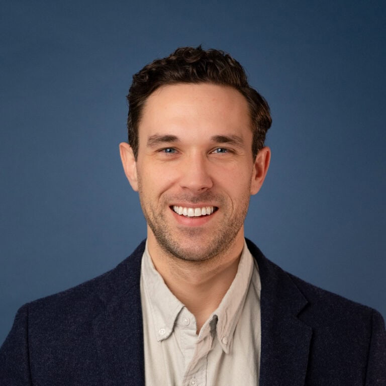 Portrait of Ben Beasley smiling in a blazer against a blue background.