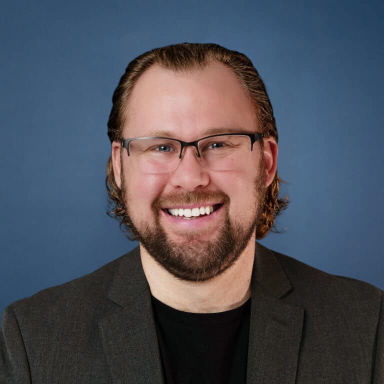 Jonathan Van Maanen smiles in a headshot with a beard, glasses, and a gray suit jacket.