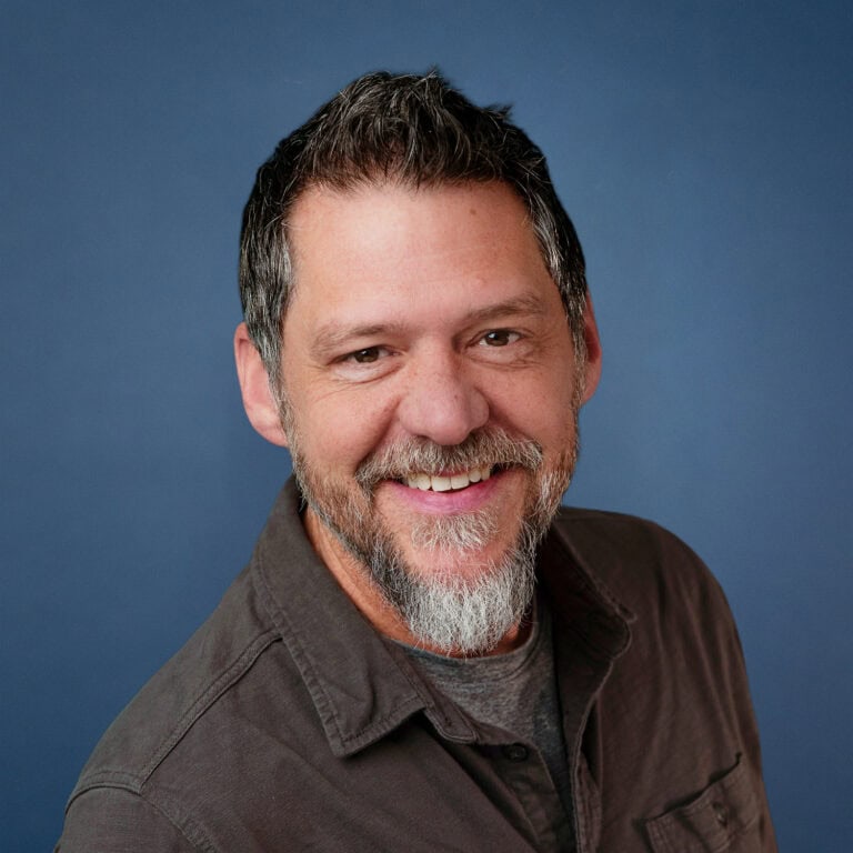 Portrait of Patrick Largen smiling with a gray beard and graying hair against a blue background.