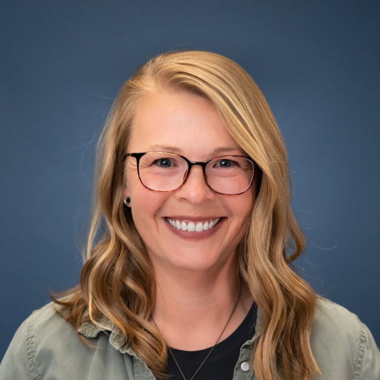 Liz Downes headshot: Smiling woman with blonde hair and glasses against a blue background.