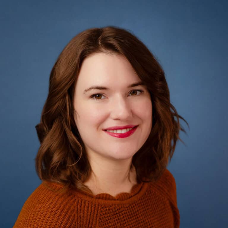 Headshot of Annica Anderson with auburn hair, wearing an orange sweater, against a blue background.