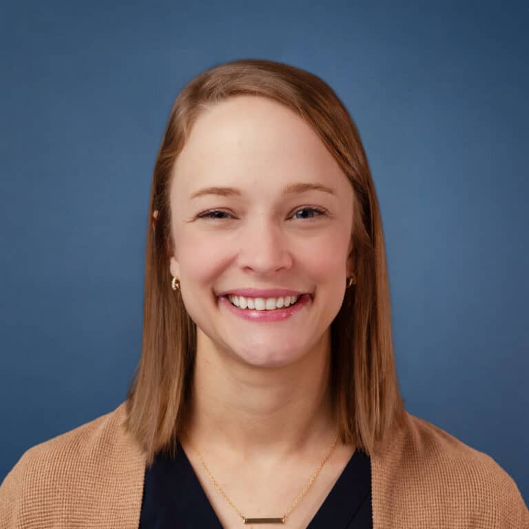 Portrait of Rachel Nesse smiling, wearing a tan cardigan and gold necklace, against a blue background.
