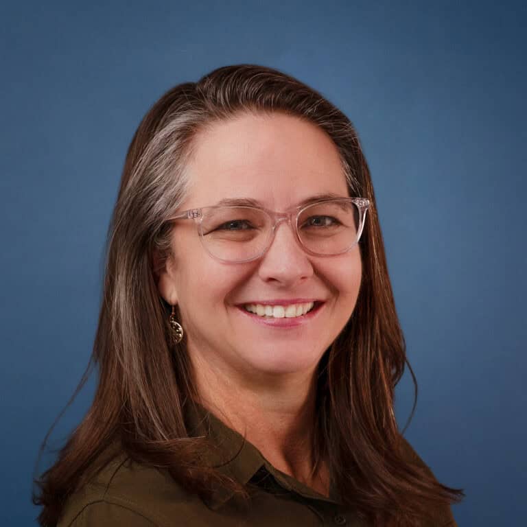 Portrait of Shanda Moon, smiling with glasses and long brown hair against a blue background.