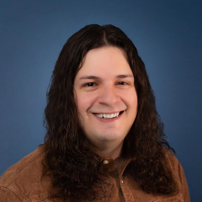 Smiling Chris Stolberg with long hair in a brown corduroy shirt against a blue background.