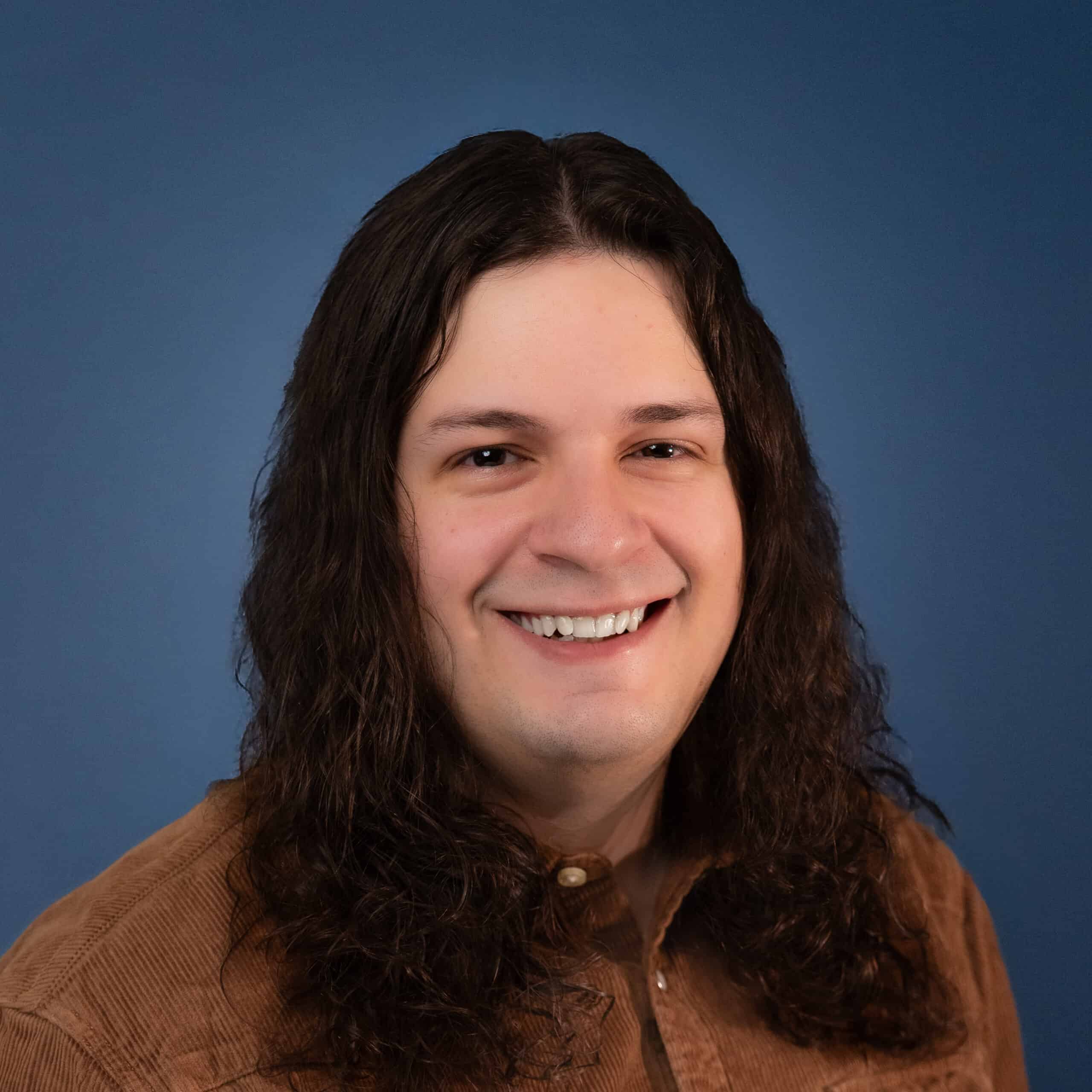 Smiling Chris Stolberg with long hair in a brown corduroy shirt against a blue background.