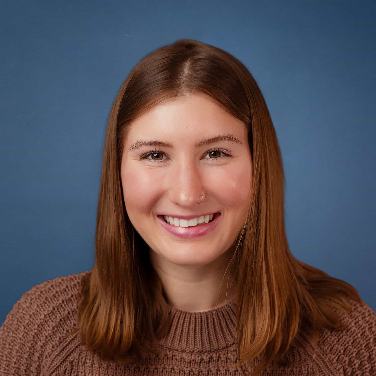 Portrait of Carlie Gregg smiling, wearing a brown sweater against a blue background.