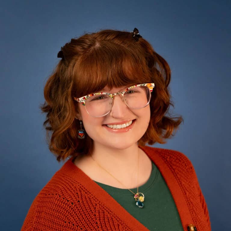 Maddie Huisman smiles, wearing colorful glasses and a cardigan.