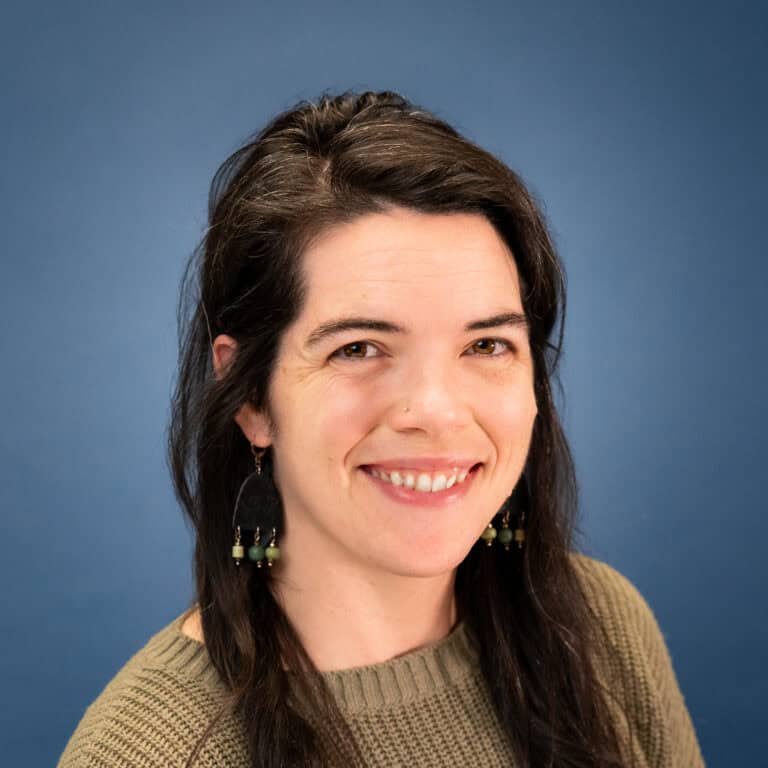 Portrait of Amanda Gibson smiling against a blue background, wearing a green sweater and earrings.