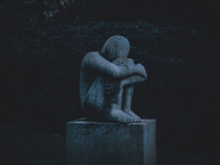 Stone statue of a person huddled in grief on a pedestal, reflecting themes of loss.