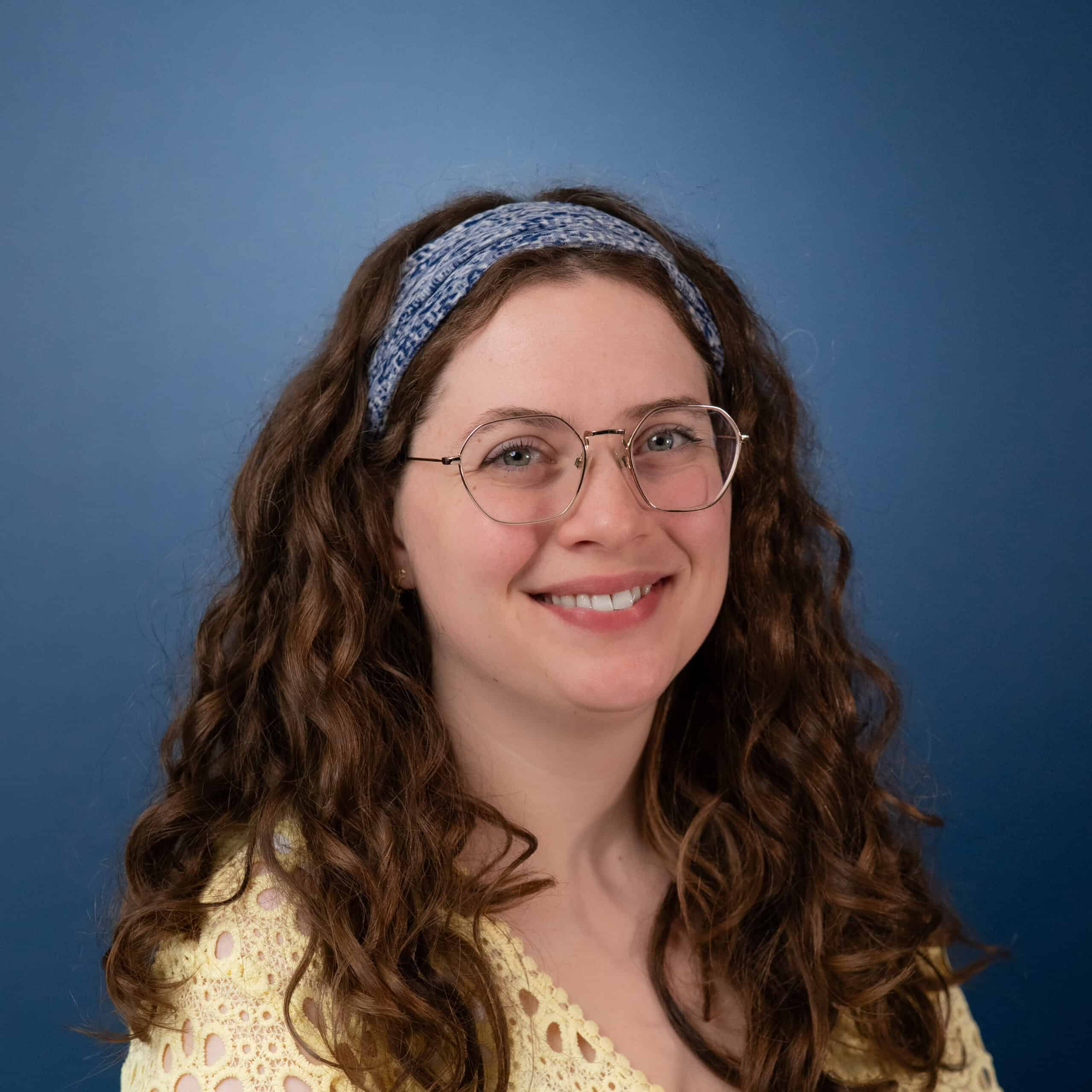 Portrait of Erin Johnson with curly hair, glasses, and a blue headband smiling.