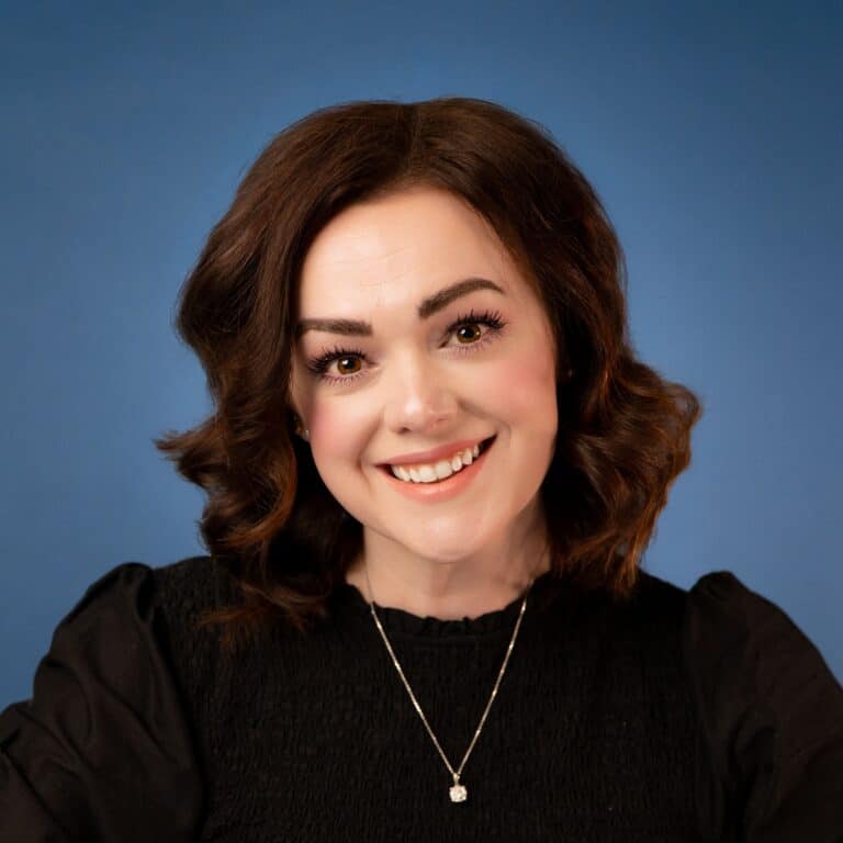 Portrait of Kristen George, smiling with dark wavy hair against a blue background.