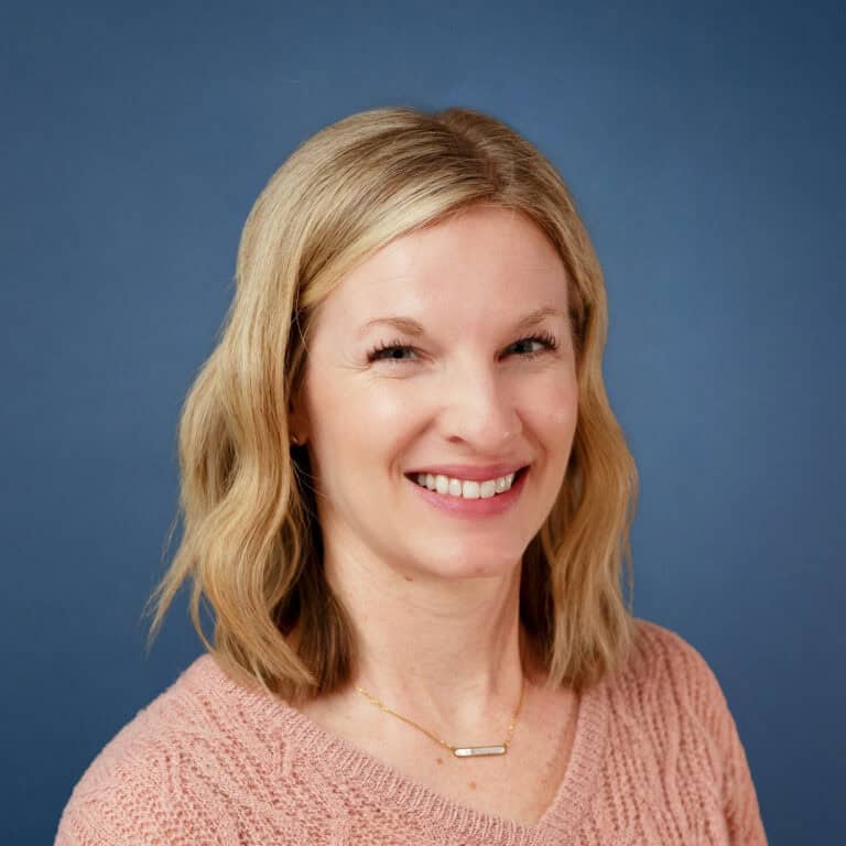 Michelle Carlson smiles in a headshot with a blue background and pink sweater.