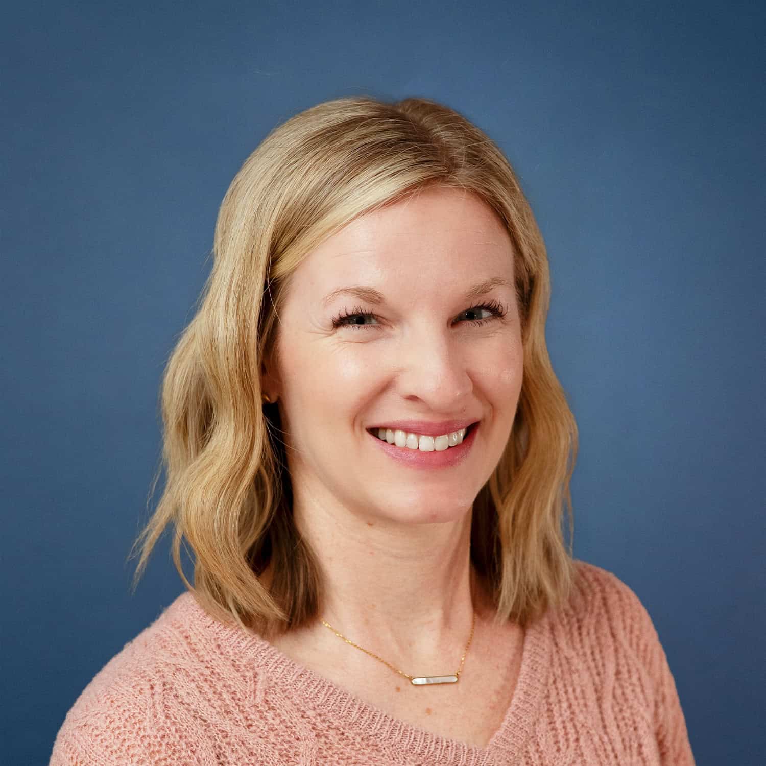 Michelle Carlson smiles in a headshot with a blue background and pink sweater.