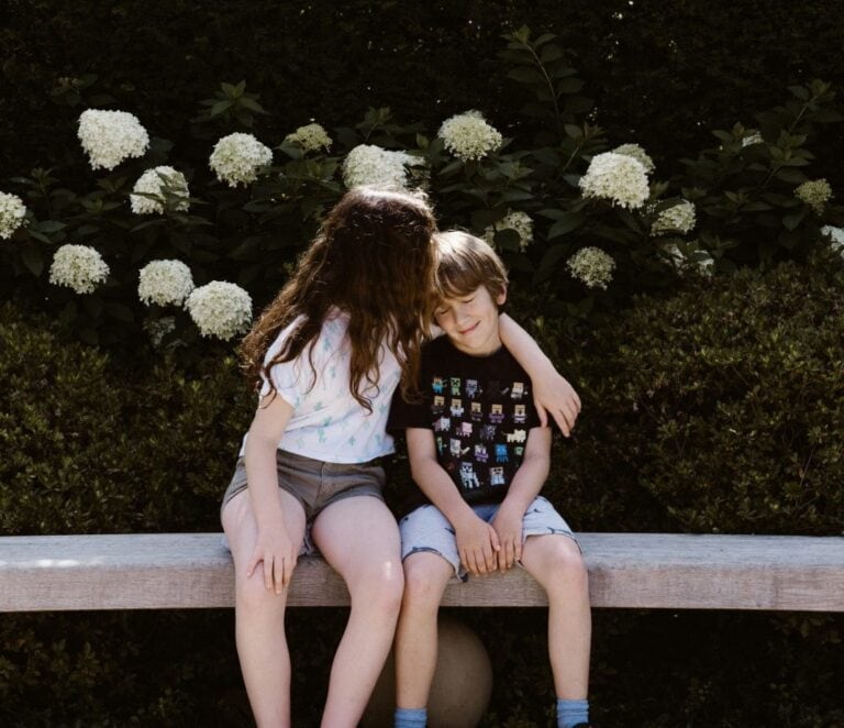 Sister hugging her brother on a bench in front of white hydrangeas.