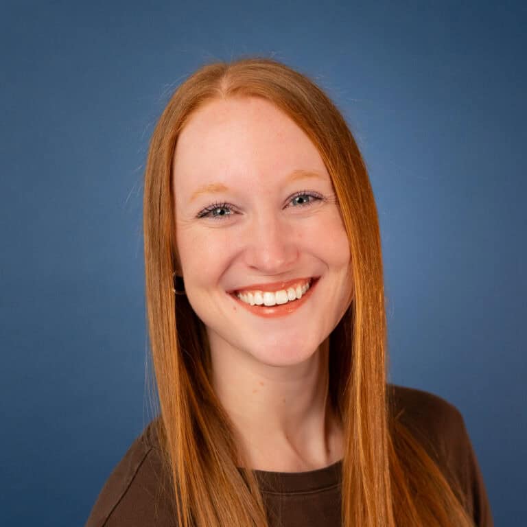 Angel Roth smiles brightly with red hair against a blue background.