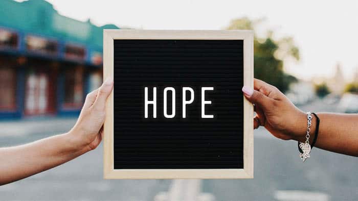 Hands holding a sign that reads "HOPE" against a blurred street backdrop.