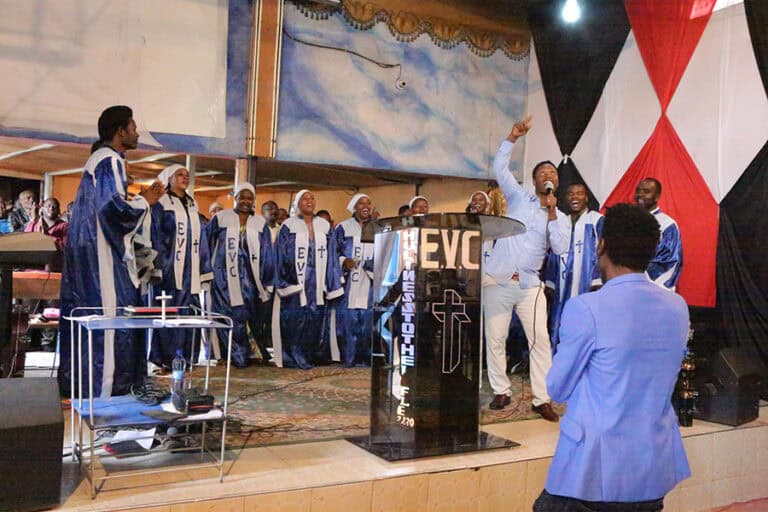 Church choir singing on stage, wearing robes. Man in blue suit kneels in foreground.