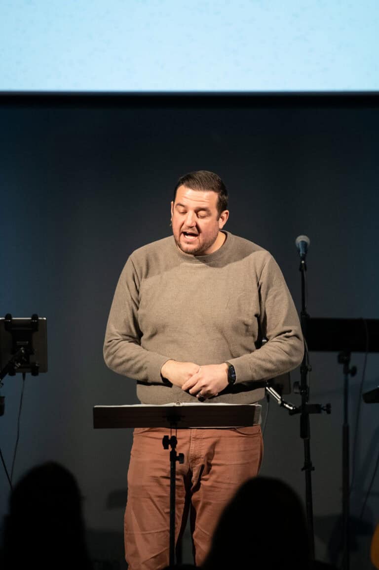 Man speaking at a podium with a microphone on stage, possibly for a podcast recording.