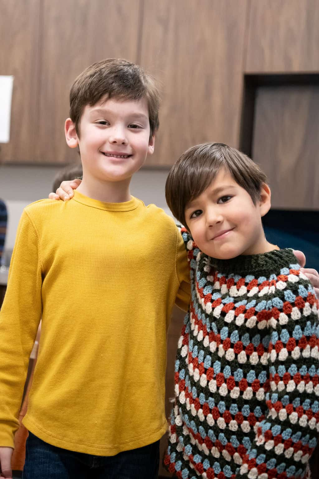 Two smiling boys, one in a yellow shirt, the other in a colorful sweater, hugging. Children's Ministries.