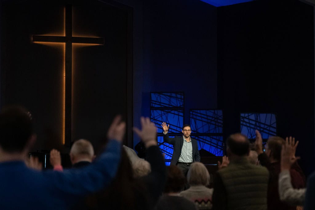 Man speaking at Leawood Campus church service with raised hands and cross