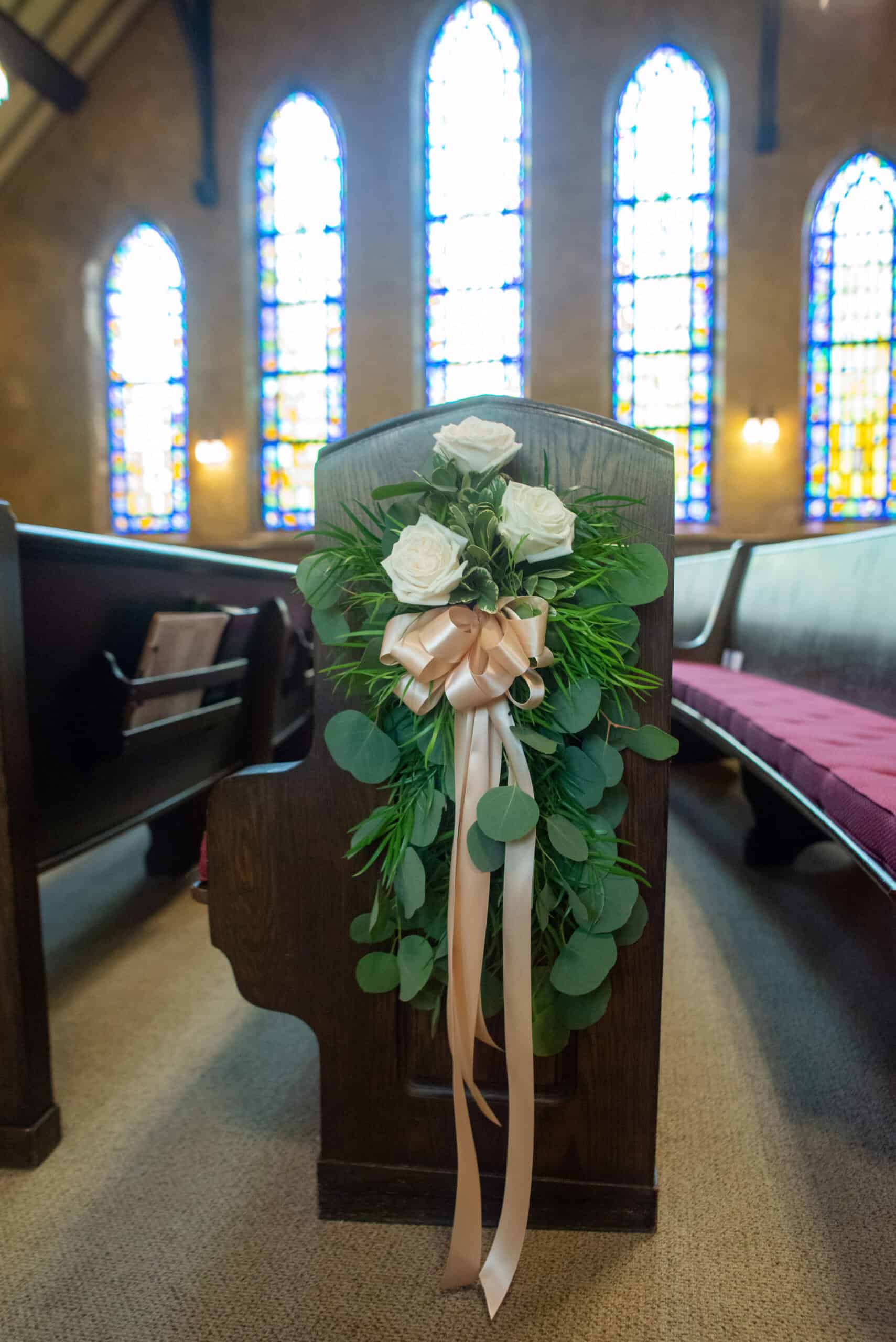 Church wedding pew decorated with white roses, greenery, and a peach ribbon, stained glass windows in background.